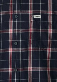 Camicia a quadri blu navy con motivo a scacchi rossi e bianchi, chiusura con bottoni, e un'etichetta Wrangler cucita sul lato sinistro. Tessuto in cotone morbido.