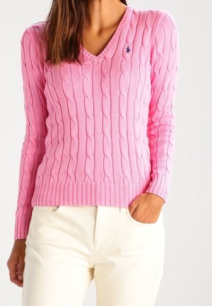 Kobieta w różowym swetrze z dzianiny warkoczowej z dekoltem w serek i jasnobeżowych spodniach, z jedną ręką w kieszeni na jednolitym tle.