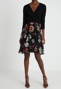 Schwarzes Kleid mit langen Ärmeln und V-Ausschnitt. Der florale Rock zeigt mehrfarbige Rosen auf schwarzem Grund. Glattes, leichtes Material.