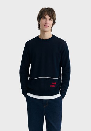 Jeune homme portant un pull noir avec deux poissons rouges et une ligne horizontale blanche, associé à un jean bleu foncé et une chemise blanche en dessous.