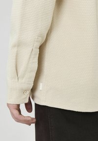 Chemise beige à texture gaufrée avec un poignet à bouton et une fente sur le côté, associée à un tissu sombre. Dispose d'une étiquette tissée simple à l'ourlet.