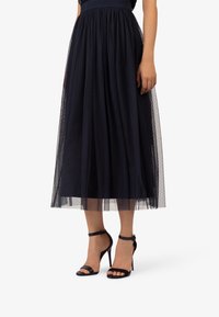 Marineblauwe midi-rok van tule met een aansluitende tailleband, voorzien van een gestippeld patroon en een gelaagde uitstraling. Gecombineerd met zwarte sandalen met hoge hak en bandjes.