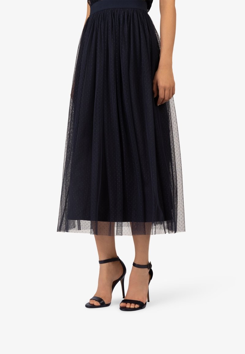Marineblauwe midi-rok van tule met een aansluitende tailleband, voorzien van een gestippeld patroon en een gelaagde uitstraling. Gecombineerd met zwarte sandalen met hoge hak en bandjes.