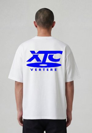Persoon met kort haar, gekleed in een wit T-shirt met grote blauwe letters "XTC" en kleinere tekst "VERTERE" op de achterkant, tegen een neutrale achtergrond.