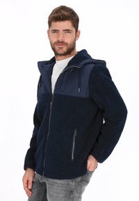 Chaqueta azul marino de forro polar con cremallera y capucha, con un panel superior liso en contraste. Cuenta con dos bolsillos laterales y un ajuste relajado.