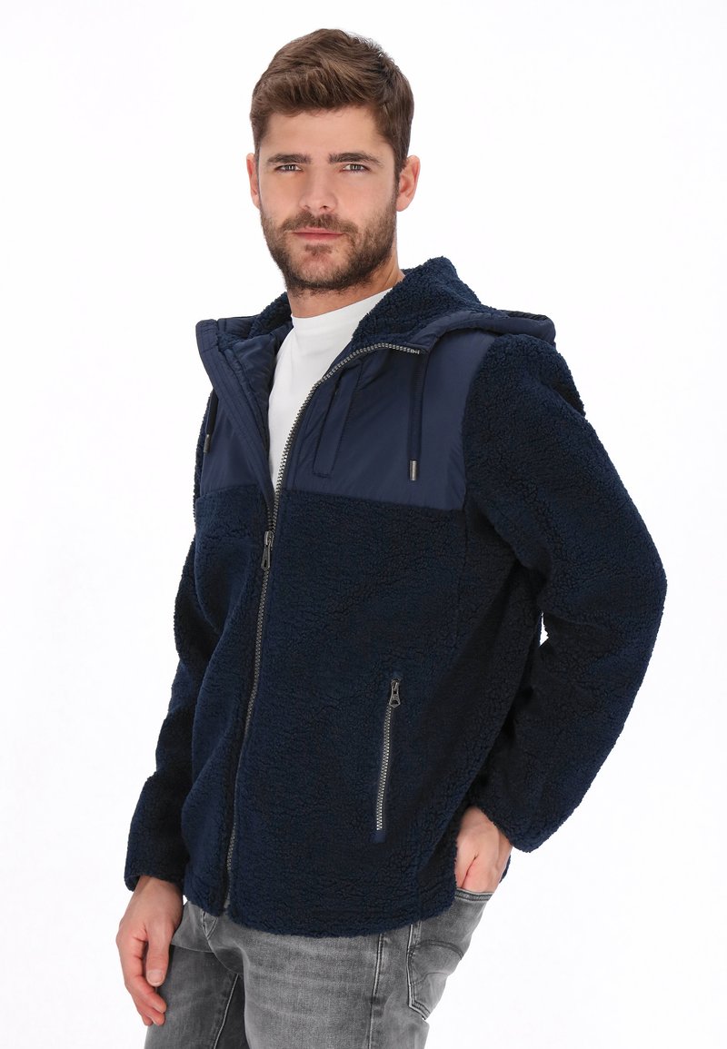 Chaqueta azul marino de forro polar con cremallera y capucha, con un panel superior liso en contraste. Cuenta con dos bolsillos laterales y un ajuste relajado.