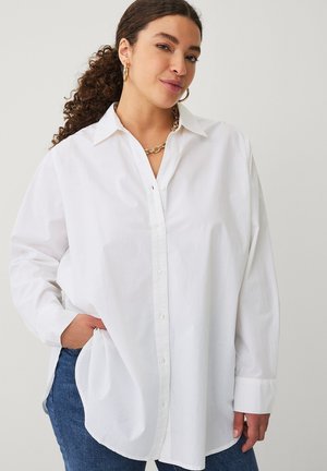 Frau mit lockigen Haaren, die goldene Creolen, eine Kettenkette, ein übergroßes weißes Hemd und blaue Jeans trägt, mit der Hand in der Tasche.