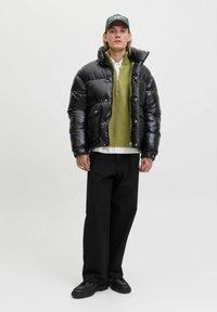 Mladostnik oblečen v črno puhovko, zeleno zip nedeljo, črne hlače, črne čevlje in črno kapo, stoji ob preprosti ozadju.