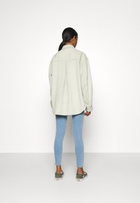 Camicia verde oversize con colletto e piega posteriore, abbinata a jeans skinny blu e scarpe slip-on verdi. Sfondo bianco uniforme.