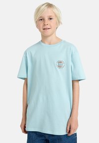 T-shirt in cotone azzurro con maniche corte, caratterizzato da un logo circolare blu e bianco sul lato sinistro del petto. Vestibilità rilassata e texture morbida.