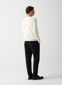 Suéter crema con un diseño simple, dobladillo acanalado, combinado con pantalones negros. El modelo está de espaldas, mostrando un estilo minimalista.