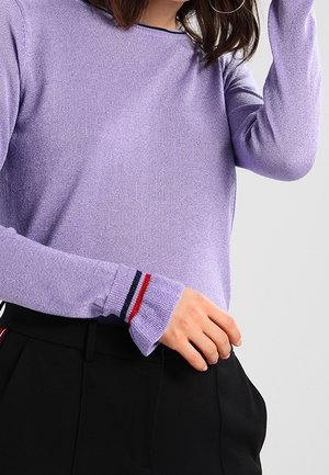 Femme portant un pull à manches longues violet clair avec des rayures rouges, blanches et bleues aux poignets et un pantalon noir sur un fond uni.