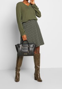 Sac à main en cuir noir avec des poignées supérieures et des accents dorés ; le modèle porte un chemisier vert et une jupe à pois, accompagnés de bottes brunes jusqu'aux genoux.