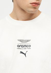 Κοντινό πλάνο ενός ατόμου που φοράει λευκό πουκάμισο με λογότυπα της Aston Martin, Aramco Formula One Team και Puma στο στήθος.