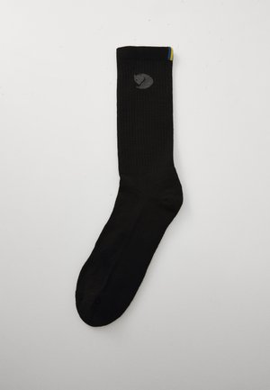 Black ribbed sock with a small gray animal logo near the top and a thin yellow and blue stripe at the cuff on a plain white background.