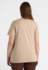 Camiseta de manga corta beige con cuello redondeado, hecha de tela suave, presenta puños enrollados y un dobladillo recto. Combinada con pantalones marrones.