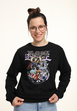 Lächelnde Frau mit Brille, die einen schwarzen Yu-Gi-Oh! GX Pullover mit Anime-Charakteren und Kreaturen trägt, posiert vor einem schlichten weißen Hintergrund.
