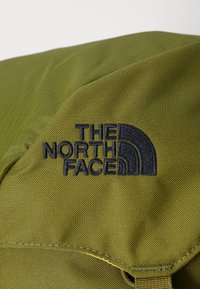 Alyvuogių žalios spalvos audinio kuprinė su tekstūruota paviršiumi, turinti juodą siuvinėtą "The North Face" logotipą ir išlenkto akcento dizainą.
