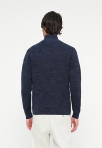 Mørkeblå strikket genser med høy krage og lange ermer. Har et teksturert mønster med ribbet kant og mansjetter.