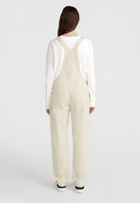 Beige cotton overalls with adjustable straps, side pockets, and a relaxed fit. Worn over a white long-sleeve shirt.