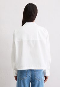 White, cropped jacket with a collared neckline, long sleeves, and button cuffs. Smooth fabric with a straight back seam and subtle detailing.