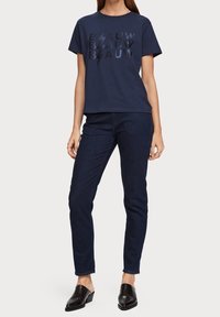 Camiseta de algodón azul marino con el texto en negrita "BLAUW" en negro y rayos. Combinada con jeans oscuros de mezclilla y zapatillas negras sin cordones.