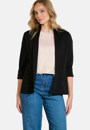 Blazer nero corto con una texture liscia, davanti aperto e due tasche frontali. Indossato sopra una maglietta chiara e abbinato a jeans blu.