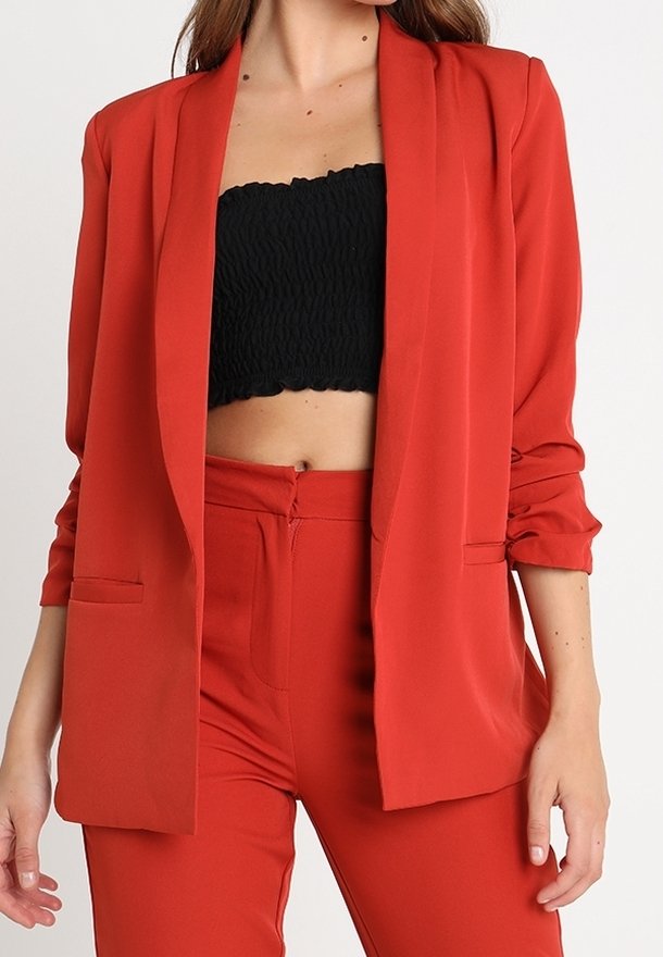 Czerwona marynarka z długimi rękawami, z wyraźnymi ramionami, dwiema przednimi kieszeniami, noszona na czarnym topie z marszczeniami, w zestawieniu z dopasowanymi czerwonymi spodniami.