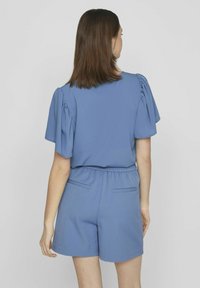 Haut bleu à manches courtes avec des épaules froncées, associé à un short bleu. Le tissu semble lisse et léger ; taille élastique avec poches arrière.