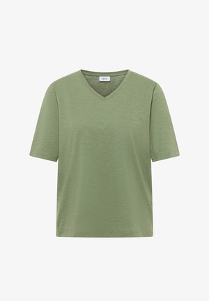 Femme aux cheveux courts et bruns portant un t-shirt vert uni à manches courtes et un jean bleu, de profil avec un fond neutre.