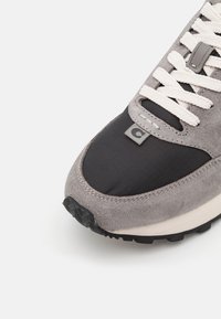 Gros plan sur l’avant d’une basket grise et noire avec des lacets blancs, texture en daim et petite étiquette logo sur fond blanc.