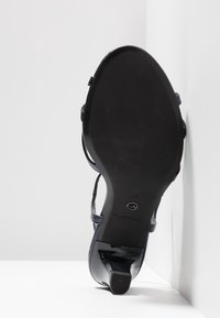 Semelle noire d'une sandale à talon haut pour femmes avec des brides, taille 37, posée verticalement contre une surface d'angle blanche.
