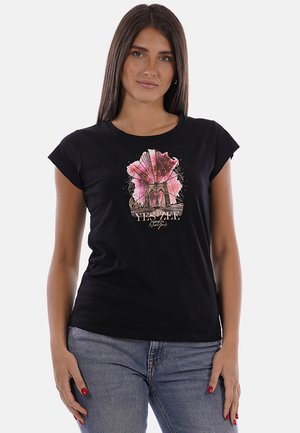 Donna con lunghi capelli castani che indossa una t-shirt nera con una grafica rosa e beige del ponte di Brooklyn e jeans blu, in piedi su sfondo bianco.