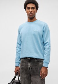 Homem com um suéter de manga longa azul claro e jeans pretos desbotados, segurando uma mala de couro preta contra um fundo branco puro.