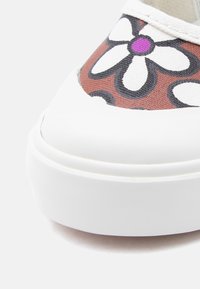 Κοντινό πλάνο μύτης λευκού αθλητικού παπουτσιού με καφέ ύφασμα που δείχνει σχέδιο λευκού και μαύρου περιγράμματος ανθέων με μωβ κέντρο.