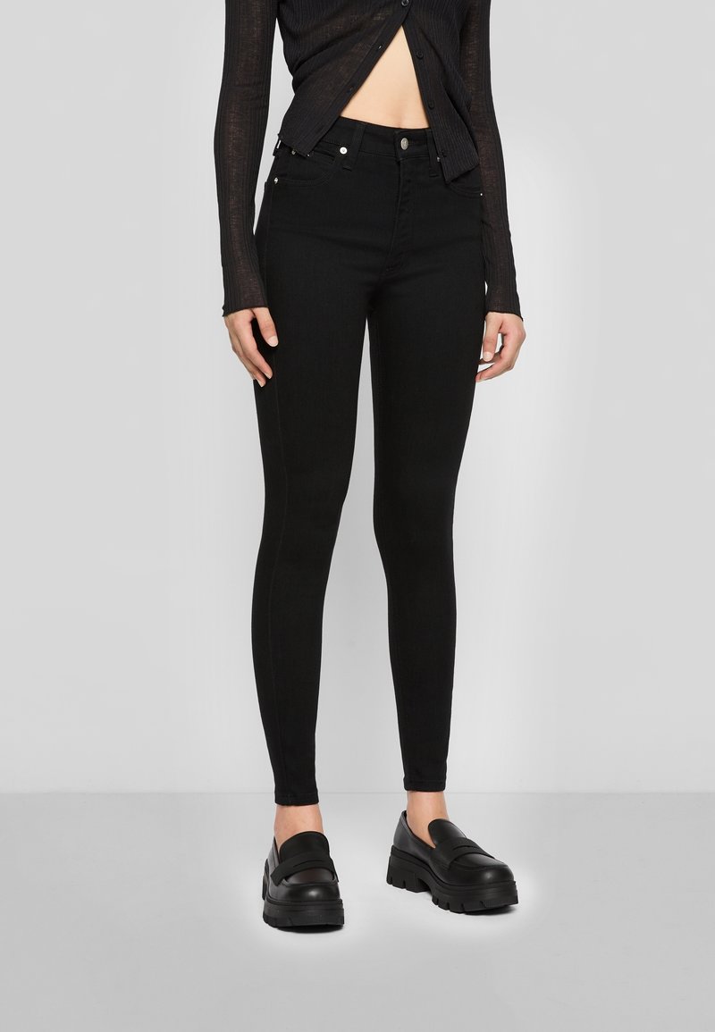 Jean noir skinny taille haute, avec une texture lisse, un design à cinq poches et une quincaillerie métallique. Associé à des chaussures noires à enfiler.
