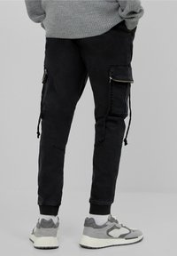 Svarta cargobyxor med blixtlåsfickor, tapered leg-design och dragsko vid fållarna, kombinerat med grå sneakers och vita ankelsockor.