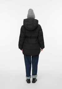 Osoba stoji okrenuta leđima, nosi crnu kapuljaču s pufastim jastučićima, sivu zimsku kapu, plave uvrnute traperice i crne cipele, nasuprot bijeloj pozadini.