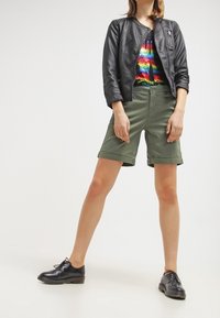 Svart läderjacka, färgglad regnbåge T-shirt, olivgröna shorts och svarta snörskor, presenterad stående, med neutral bakgrund.