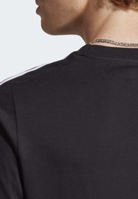 T-shirt nero con scollatura rotonda e texture morbida, con dettagli a strisce bianche lungo le spalle e una collana d'argento visibile.