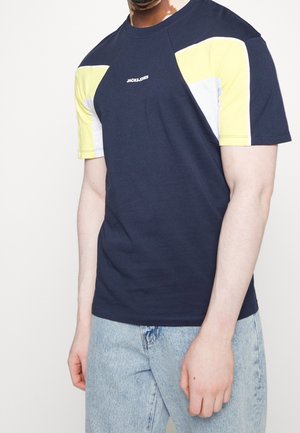 Homme portant un t-shirt bleu marine avec des panneaux d'épaule jaunes et blancs et le logo "JACK&JONES", assorti à un jean bleu clair.