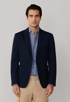 Hombre con blazer azul marino sobre camisa azul de botones y pantalones beige, de pie contra un fondo liso gris claro.