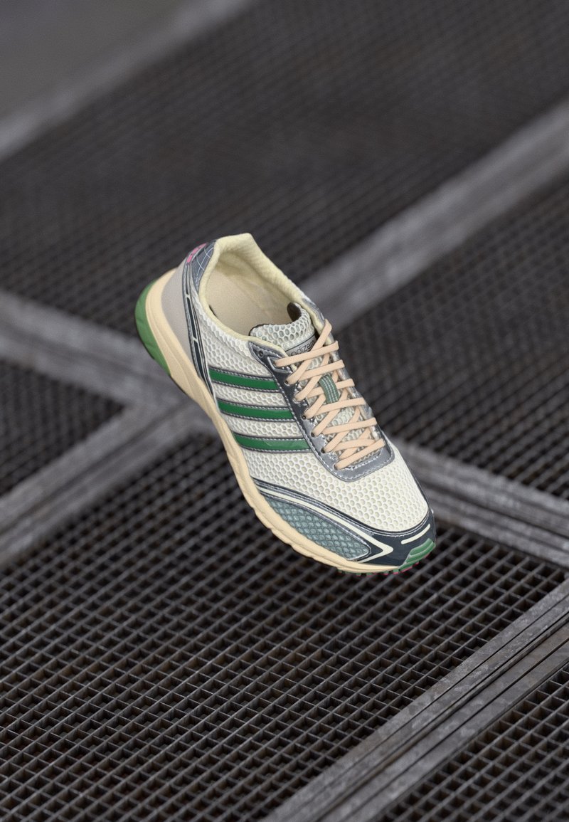 Sneaker bianca e verde in rete con lacci beige che fluttua sopra un pavimento in griglia metallica scura.