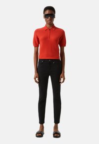 Červené polo tričko s krátkými rukávy a zipem u krku, doplněné černými úzkými džíny a černými sandály. Vzhled doplňují sluneční brýle.