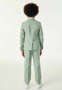 Completo verde menta con finitura testurizzata, dotato di una fessura singola sul retro, polsini abbottonati e pantaloni abbinati. Camicia bianca a maniche lunghe visibile.