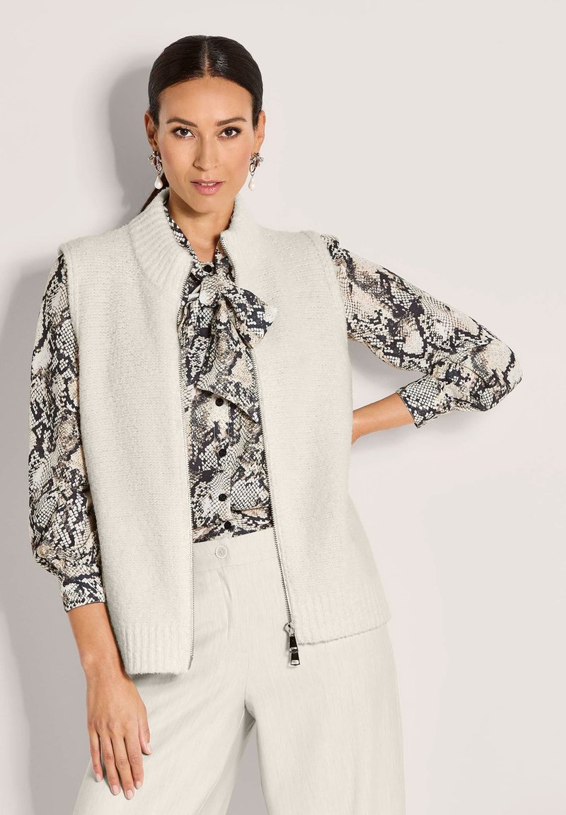 Frau mit dunklem Haar, die Perlenohrringe, eine Schlangehaut-blus mit Schleife, eine weiße Strickweste mit Reißverschluss und weiße Hosen trägt, vor einem einfarbigen Hintergrund.