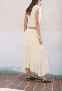 Ensemble en dentelle composé d'un haut court ajusté et d'une longue jupe dans un jaune doux. La tenue possède un ourlet fluide et de subtils motifs floraux. Talons ouverts marron.