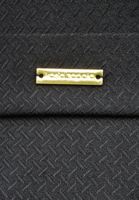 Czarna tkanina o fakturze z wzorem w rybią ość. Do tkaniny przymocowany jest prostokątny złoty tag z logo, wygrawerowany napisem "jesie zapacie."