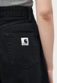 Μαύρο τζιν παντελόνι με πίσω τσέπη που δείχνει λευκό λογότυπο Carhartt, φορεμένο από άτομο με γκρι τοπ, με εμφανές χέρι στο πλάι.