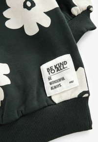 Zielona bluza z dużym białym wzorem kwiatowym, ściągacze w rękawach oraz naszywka z tekstem "BĄDŹ DOBRY DLA WSZYSTKICH. BĄDŹ ZAWSZE WSPANIAŁY."